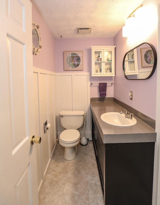 half bath with a textured ceiling, visible vents, vanity, a wainscoted wall, and toilet