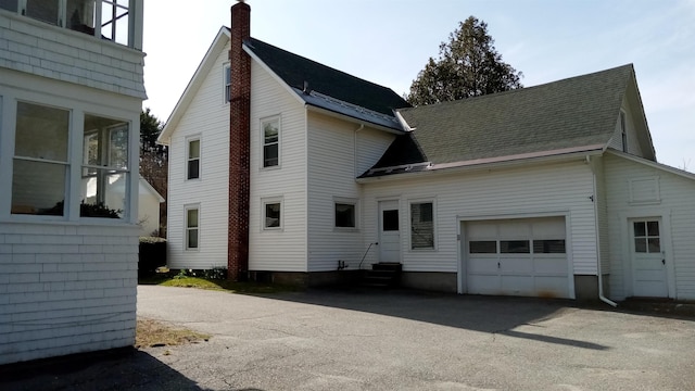 back of house featuring a shingled roof, an attached garage, entry steps, a chimney, and driveway