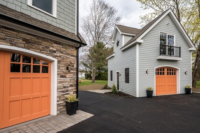 view of side of property featuring a balcony, stone siding, a shingled roof, an attached garage, and driveway