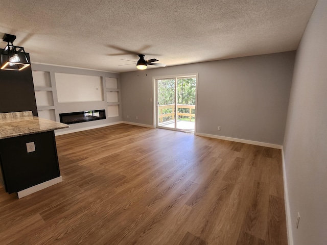 unfurnished living room with ceiling fan, a textured ceiling, a glass covered fireplace, baseboards, and wood finished floors