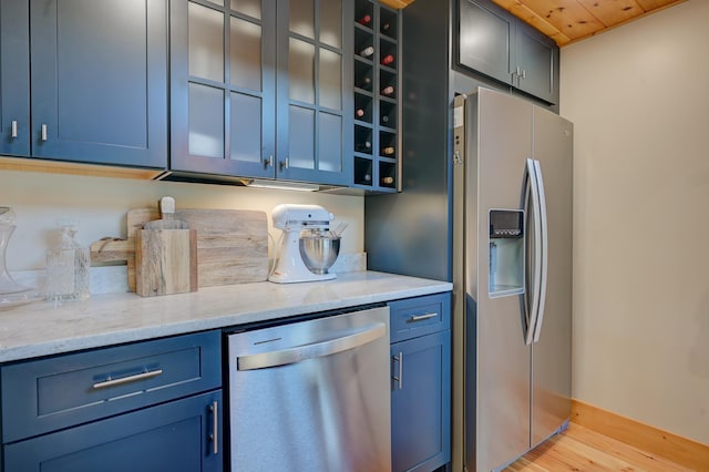 kitchen featuring appliances with stainless steel finishes, light stone counters, light wood finished floors, blue cabinetry, and glass insert cabinets