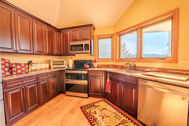 kitchen with light stone countertops, appliances with stainless steel finishes, a sink, light wood finished floors, and vaulted ceiling