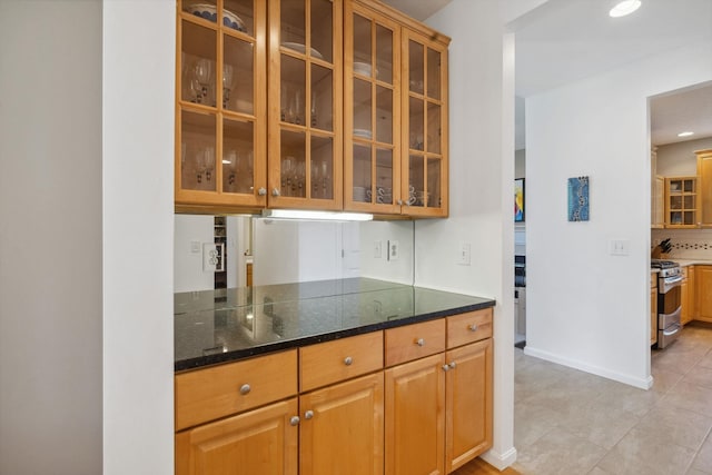 kitchen featuring gas range, baseboards, dark stone countertops, glass insert cabinets, and light tile patterned flooring