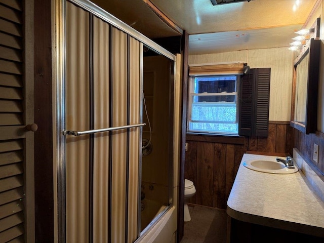bathroom with vanity, wood walls, wainscoting, toilet, and bath / shower combo with glass door