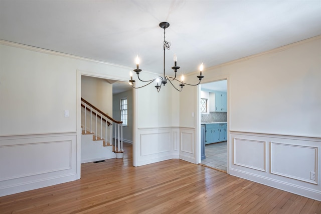 unfurnished dining area featuring wainscoting, light wood finished floors, a notable chandelier, a decorative wall, and stairs