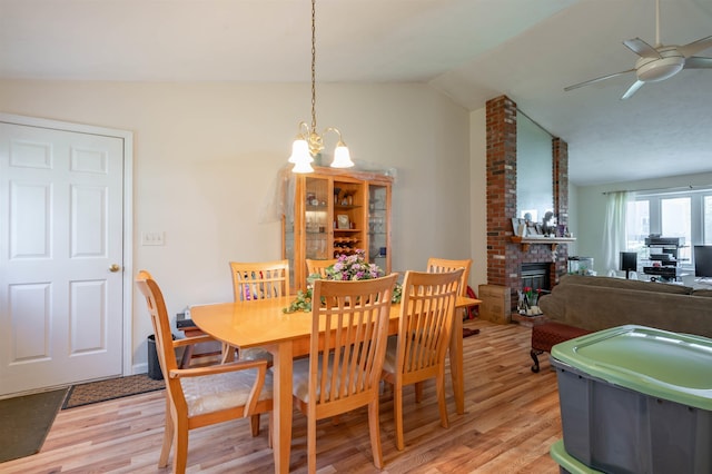 dining area featuring light wood-style flooring, ceiling fan with notable chandelier, vaulted ceiling, and a fireplace