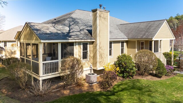 rear view of property with cooling unit, a chimney, a sunroom, a shingled roof, and a lawn