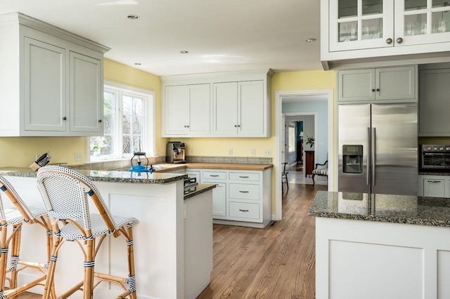 kitchen with stainless steel refrigerator with ice dispenser, a peninsula, dark stone countertops, light wood finished floors, and glass insert cabinets