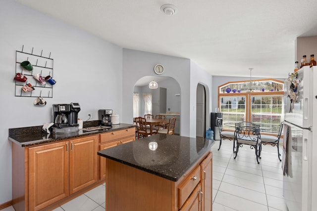 kitchen featuring freestanding refrigerator, light tile patterned flooring, a center island, dark stone counters, and arched walkways