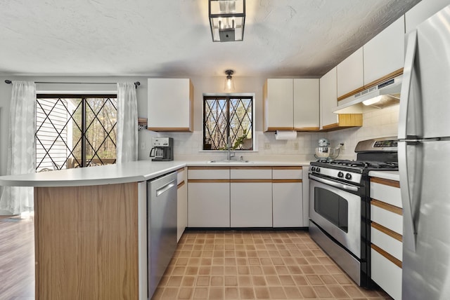 kitchen with a sink, light countertops, a peninsula, under cabinet range hood, and stainless steel appliances