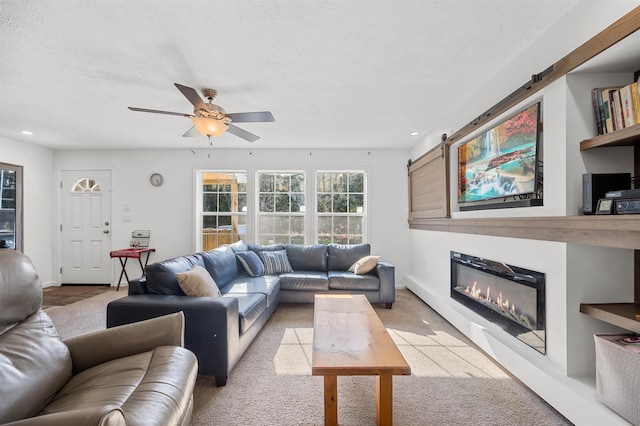 living room featuring a ceiling fan, light colored carpet, a barn door, a glass covered fireplace, and recessed lighting