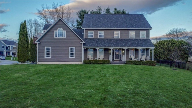 traditional-style house featuring fence, covered porch, a shingled roof, and a yard