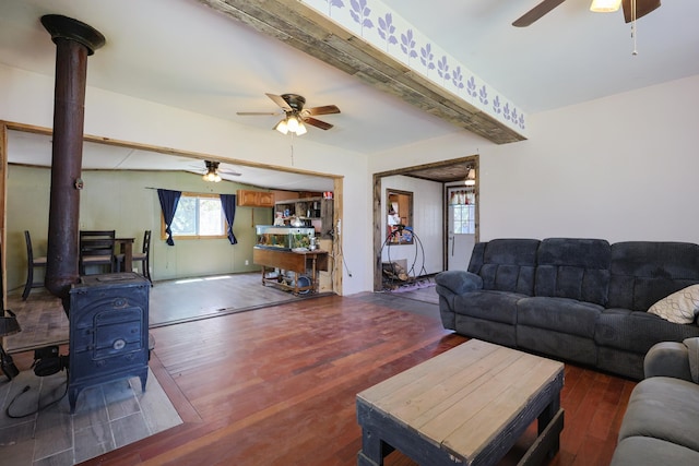living area featuring a ceiling fan, beam ceiling, wood finished floors, and a wood stove