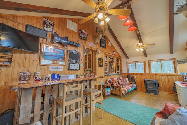 living room featuring visible vents, a wood stove, ceiling fan, wooden walls, and lofted ceiling with beams