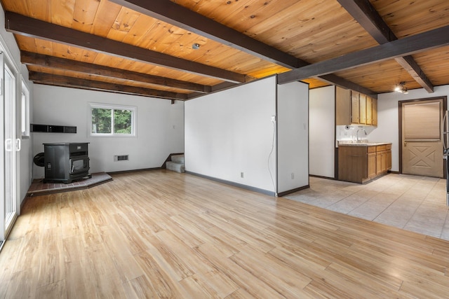 unfurnished living room featuring a wood stove, beamed ceiling, baseboards, a sink, and light wood finished floors