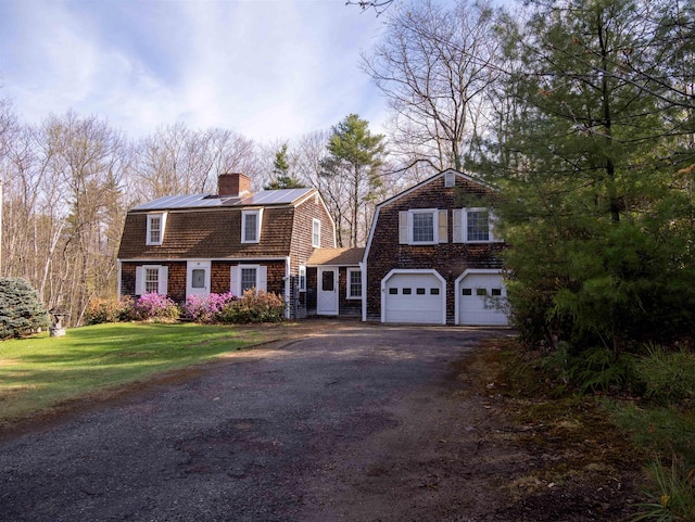 colonial inspired home with aphalt driveway, a gambrel roof, a garage, solar panels, and a chimney