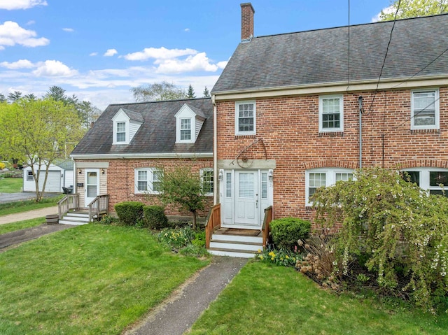 view of front facade featuring brick siding, a shingled roof, a front lawn, and a chimney