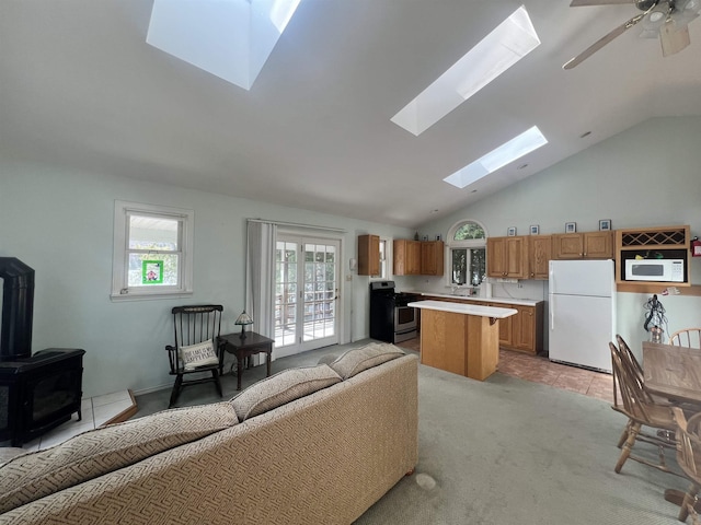 living room featuring a wood stove, a ceiling fan, vaulted ceiling with skylight, and light colored carpet