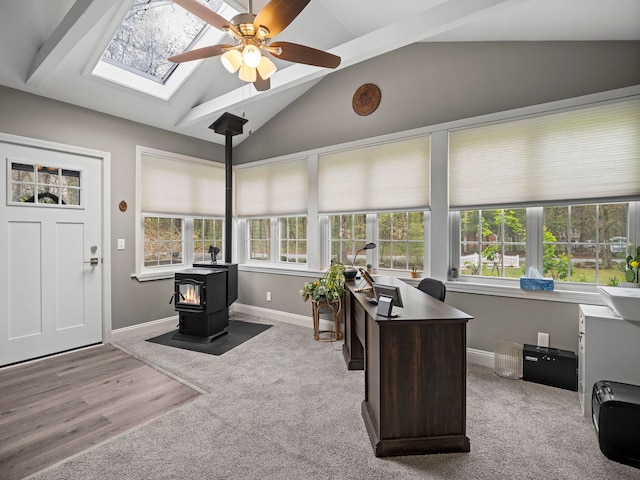 carpeted home office featuring a wood stove, lofted ceiling with skylight, baseboards, and a ceiling fan