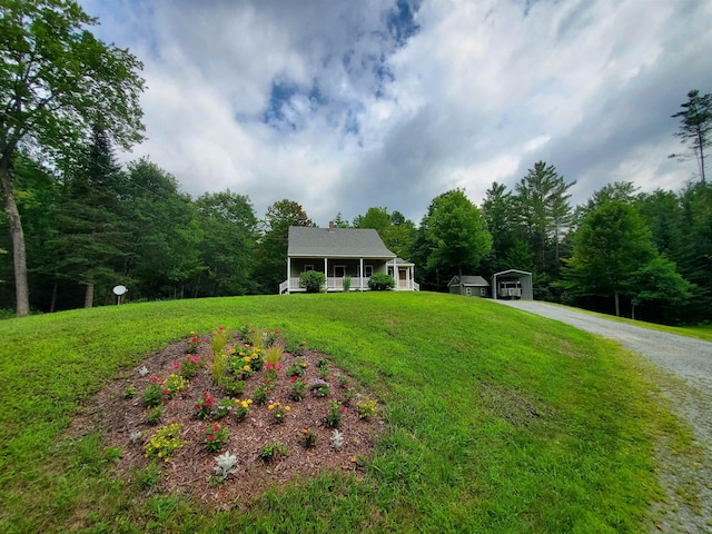 view of green lawn with a detached carport, gravel driveway, covered porch, a wooded view, and an outdoor structure