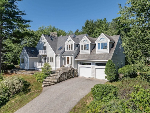 shingle-style home featuring asphalt driveway, a balcony, a chimney, a shingled roof, and stairway