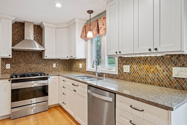 kitchen with stainless steel appliances, wall chimney range hood, a sink, light stone counters, and white cabinets