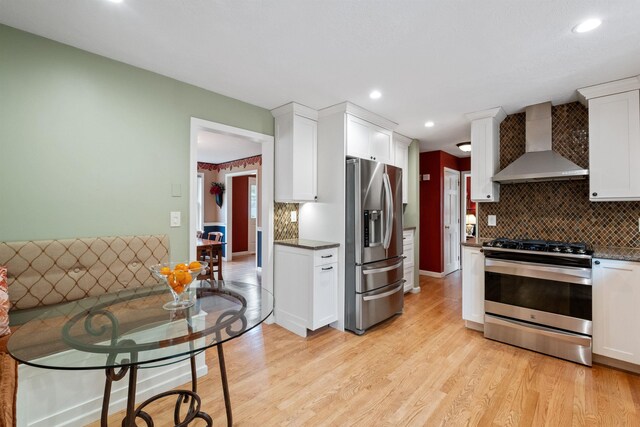 kitchen featuring appliances with stainless steel finishes, wall chimney range hood, light wood-type flooring, decorative backsplash, and white cabinets