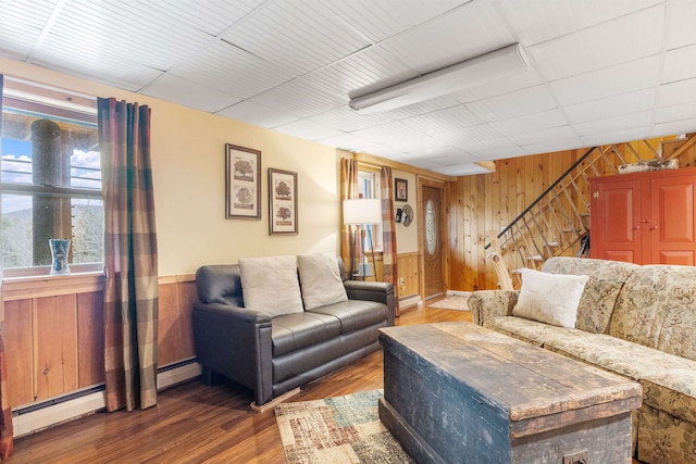 living room featuring wooden walls, stairs, wood finished floors, baseboard heating, and wainscoting