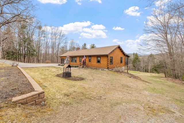 cabin with log siding, concrete driveway, a front lawn, and a chimney