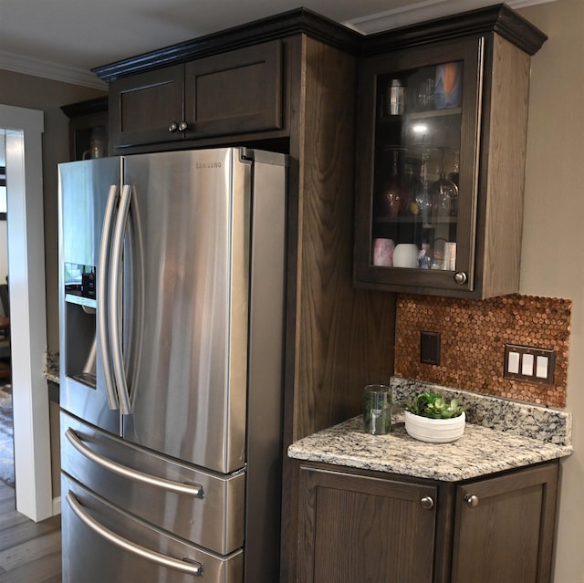 kitchen featuring stainless steel refrigerator with ice dispenser, dark brown cabinetry, glass insert cabinets, ornamental molding, and decorative backsplash