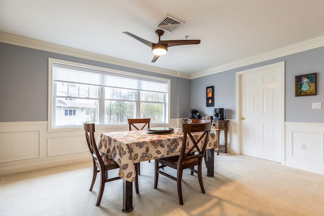 dining room featuring wainscoting, light carpet, crown molding, a ceiling fan, and a decorative wall