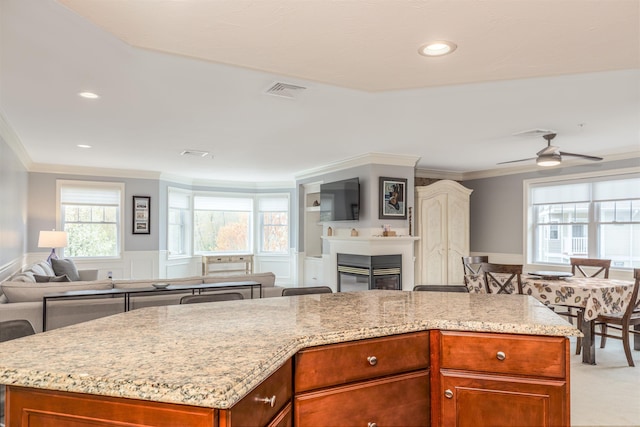 kitchen with open floor plan, ornamental molding, light stone counters, wainscoting, and a glass covered fireplace