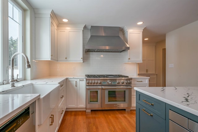 kitchen with wall chimney range hood, appliances with stainless steel finishes, white cabinets, light wood finished floors, and recessed lighting