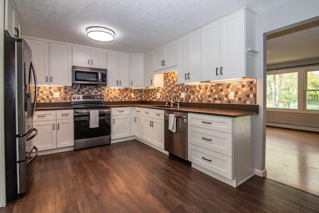 kitchen with stainless steel appliances, dark countertops, a sink, a baseboard radiator, and dark wood-type flooring