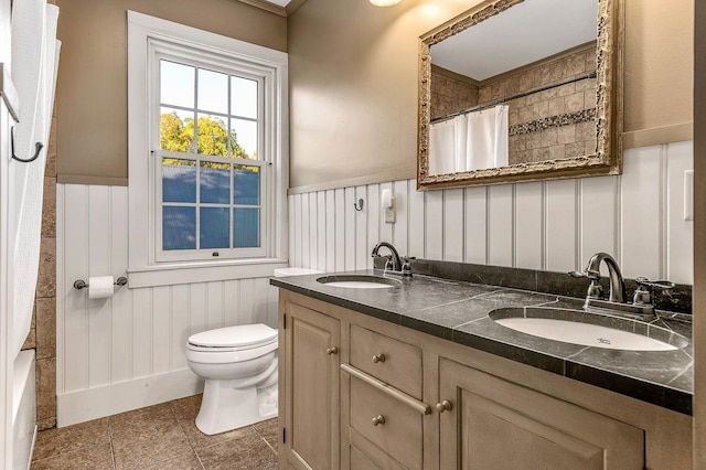 bathroom featuring wainscoting, toilet, tile patterned floors, double vanity, and a decorative wall