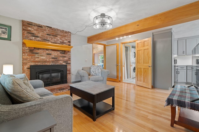 living area featuring stacked washer / drying machine, light wood-type flooring, a brick fireplace, a chandelier, and beamed ceiling