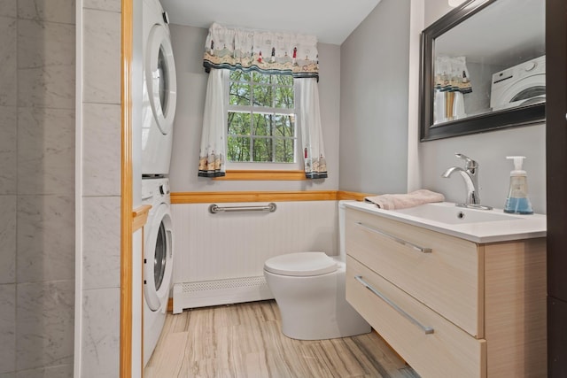 bathroom with stacked washer and clothes dryer, a baseboard radiator, toilet, a wainscoted wall, and vanity