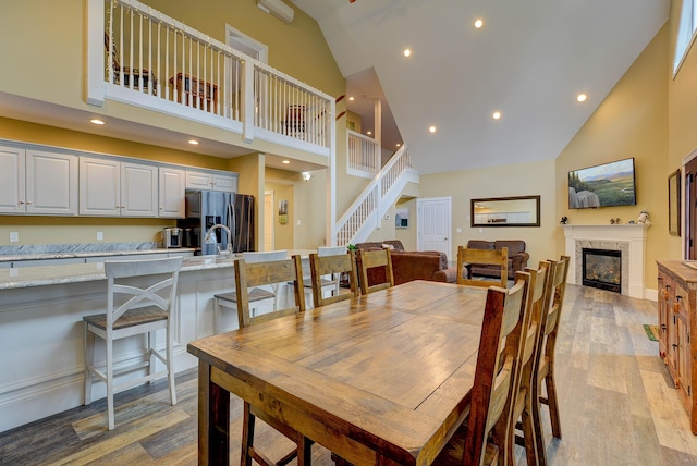 dining room featuring high vaulted ceiling, light wood-style flooring, a glass covered fireplace, recessed lighting, and stairs