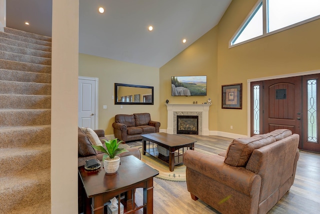 living room with high vaulted ceiling, light wood-style floors, stairs, a glass covered fireplace, and baseboards