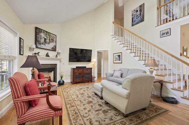 living room with stairs, wood finished floors, high vaulted ceiling, a glass covered fireplace, and baseboards