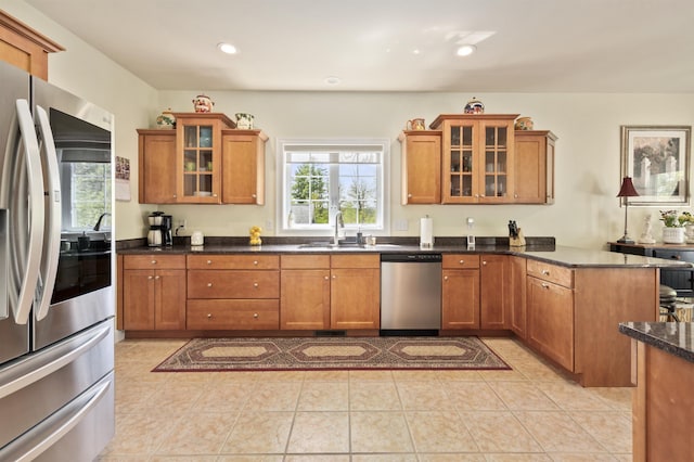 kitchen featuring appliances with stainless steel finishes, a sink, brown cabinetry, a peninsula, and glass insert cabinets