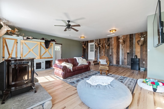 living room with wood finished floors, wood walls, a ceiling fan, and a fireplace with raised hearth
