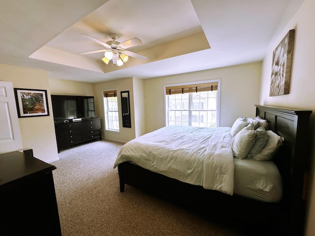 bedroom with a raised ceiling, light colored carpet, multiple windows, and ceiling fan