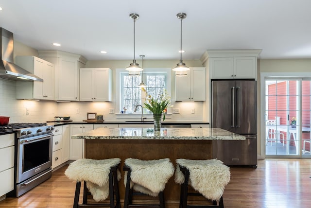 kitchen with stainless steel appliances, wall chimney exhaust hood, a sink, wood finished floors, and a center island