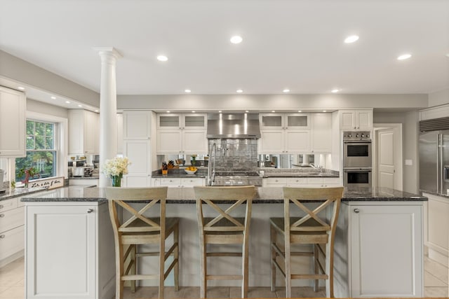 34 Orchard Hill Rd, Greenland, NH 03840 - kitchen featuring wall chimney range hood, ornate columns, a large island with sink, stainless steel double oven, and white cabinets