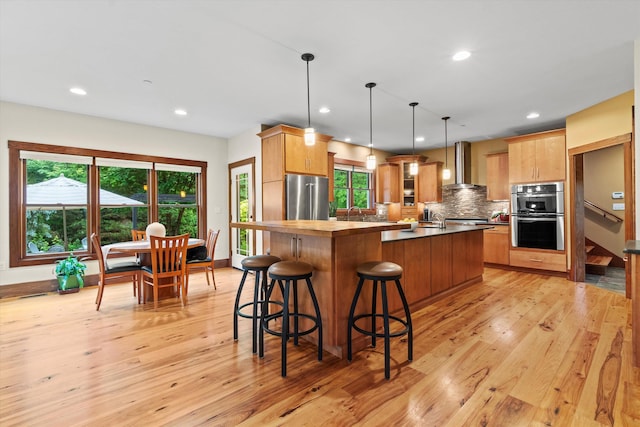 10 Benjamins Way, Hollis, NH 03049 - kitchen featuring wall chimney exhaust hood, stainless steel appliances, light wood-style floors, tasteful backsplash, and a kitchen island with sink