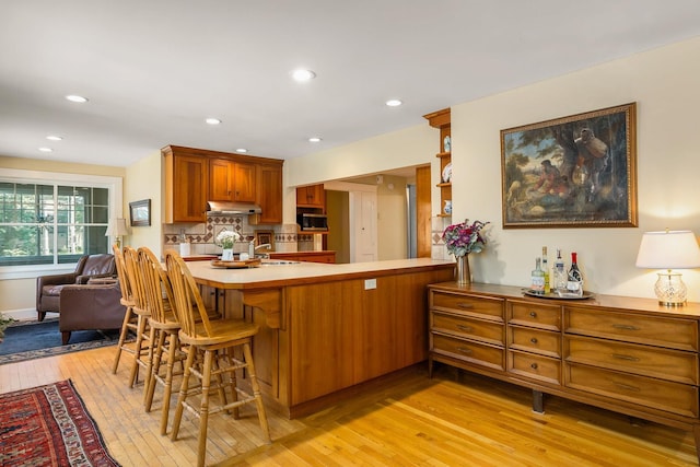 96 Madbury Rd, Durham, NH 03824 - kitchen featuring under cabinet range hood, light wood-style floors, brown cabinetry, tasteful backsplash, and a breakfast bar area