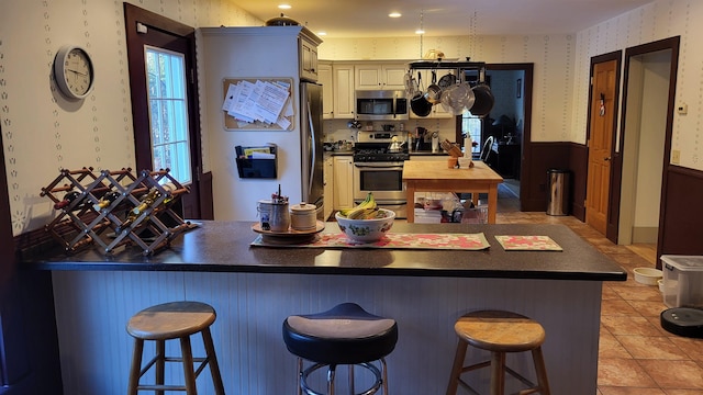 987 Dunbar Hill Rd, Grantham, NH 03753 - kitchen with stainless steel appliances, wallpapered walls, a wainscoted wall, dark countertops, and a breakfast bar