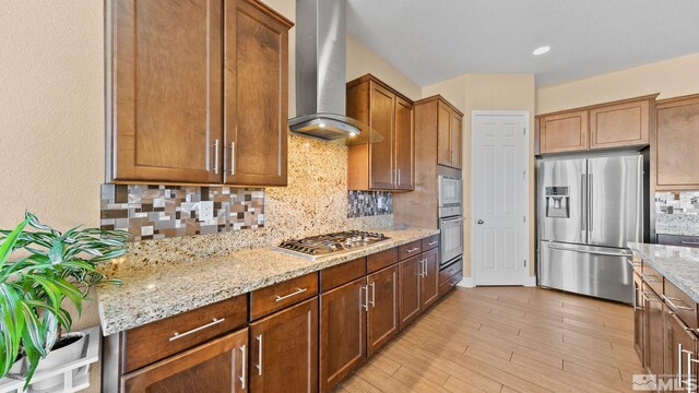 11725 Paradise View Dr, Sparks, NV 89441 - kitchen featuring appliances with stainless steel finishes, wall chimney range hood, light stone counters, decorative backsplash, and light wood-type flooring