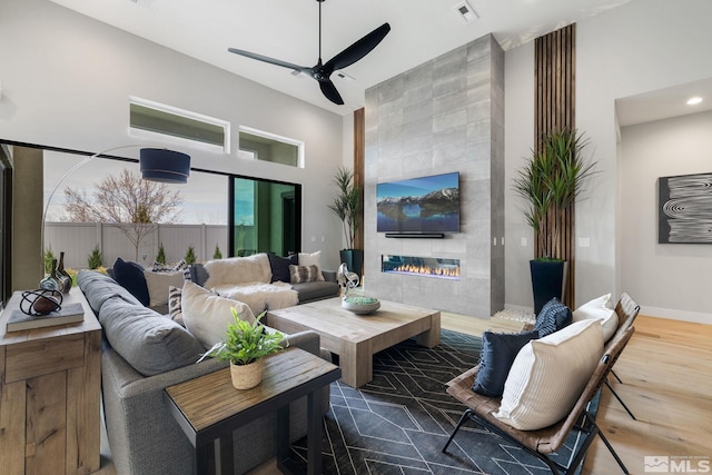 living area with ceiling fan, wood finished floors, a high ceiling, a tiled fireplace, and baseboards